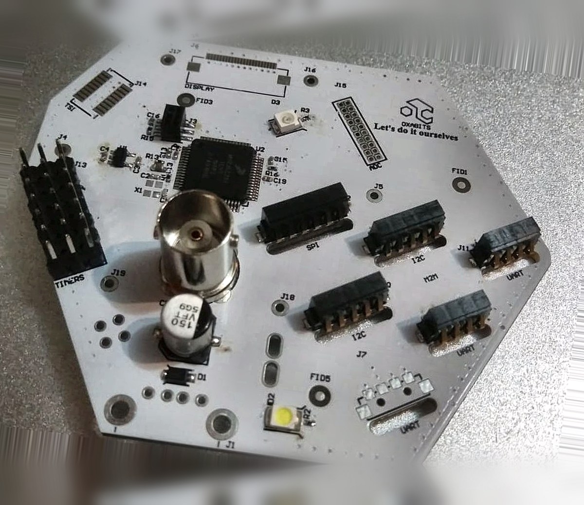 Placa electrónica hexagonal blanca diseñada por OXABITS, con microcontrolador, conector BNC y puertos SPI, I2C y UART